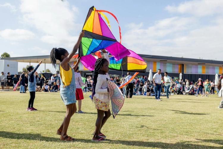 two children holding a large colourful kite with a building and people in background